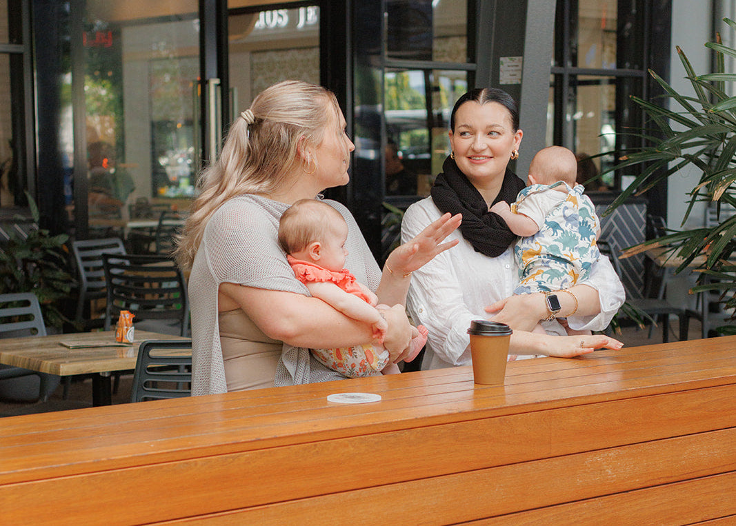 Two women with babies sitting at a wooden table in an outdoor cafe setting wearing their cowries.